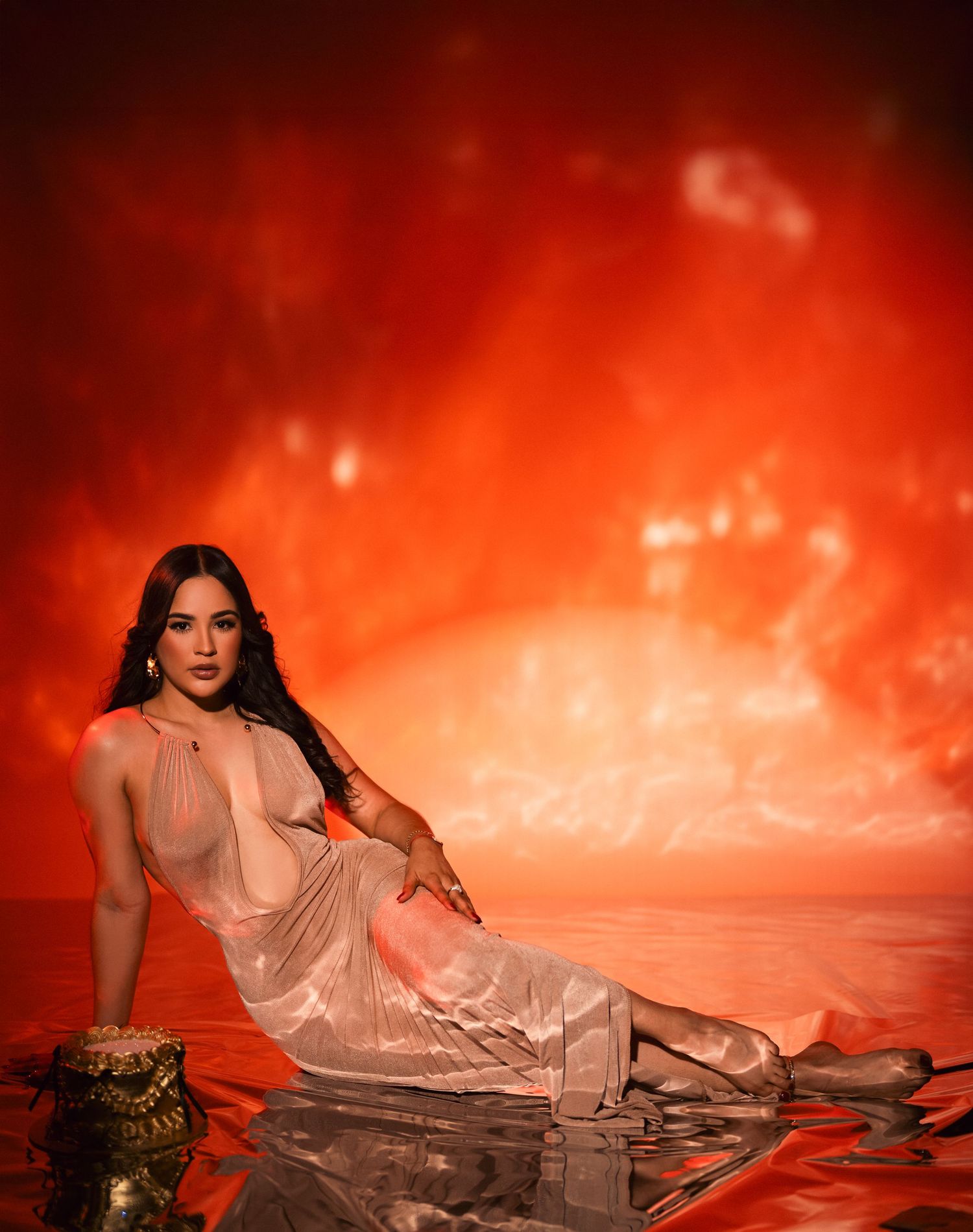 A person in a flowing beige dress poses elegantly against a dramatic red and orange sunset backdrop near water.