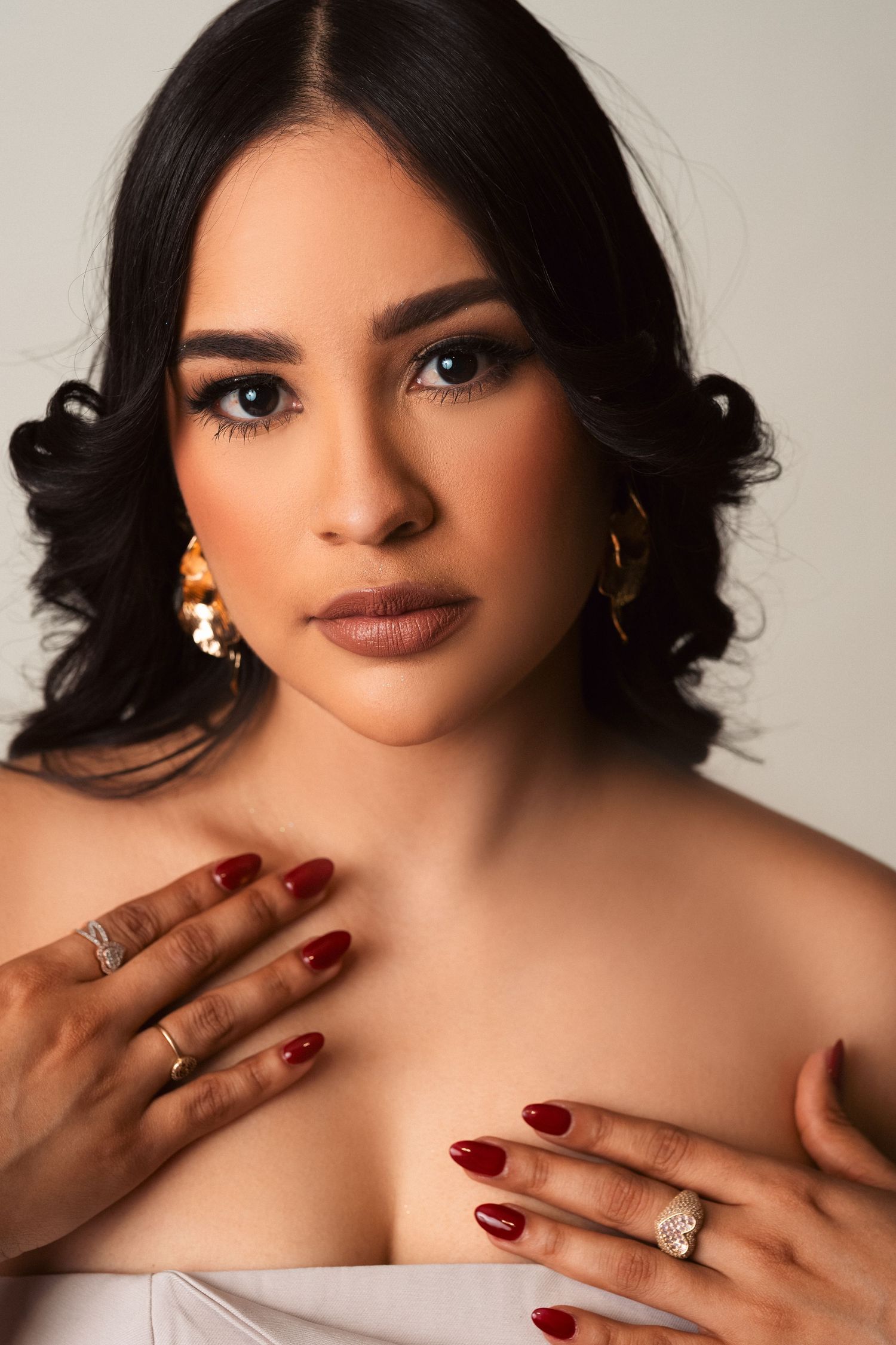 Glamour portrait featuring manicured hands with red nails, dark wavy hair, and elegant jewelry in soft studio lighting.