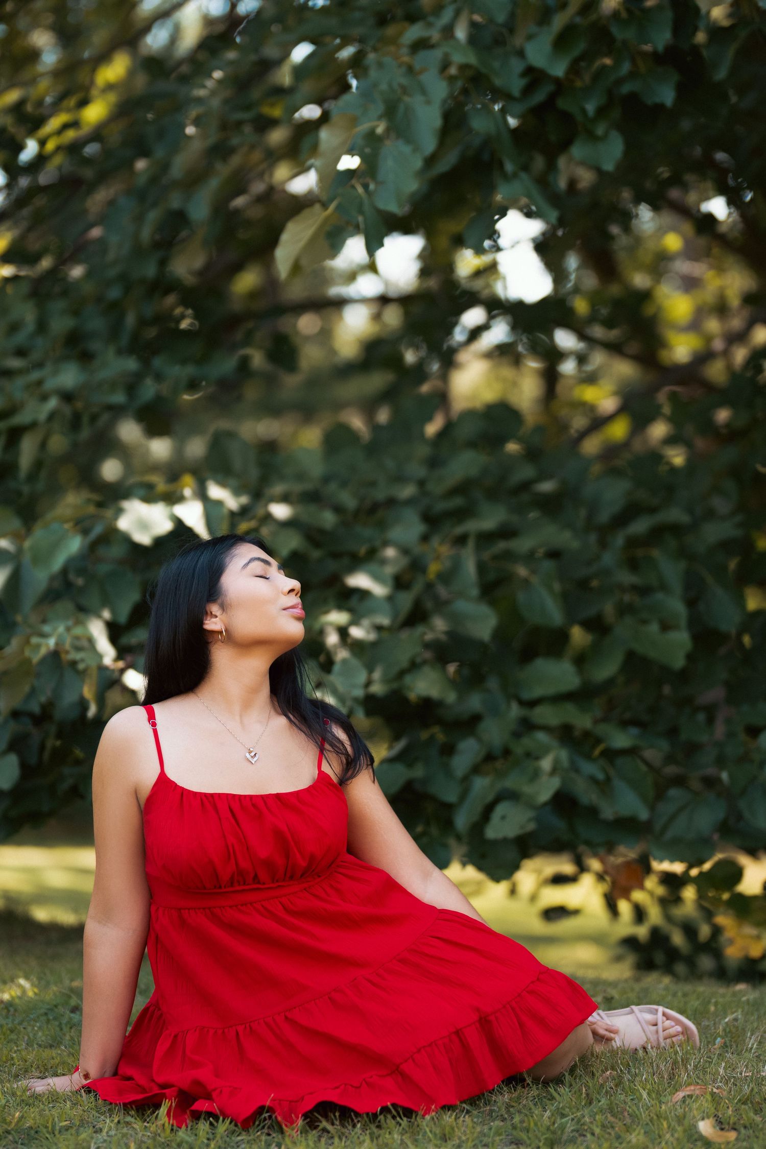 A person in a flowing red dress lies gracefully on green grass amidst autumn foliage in an outdoor setting.