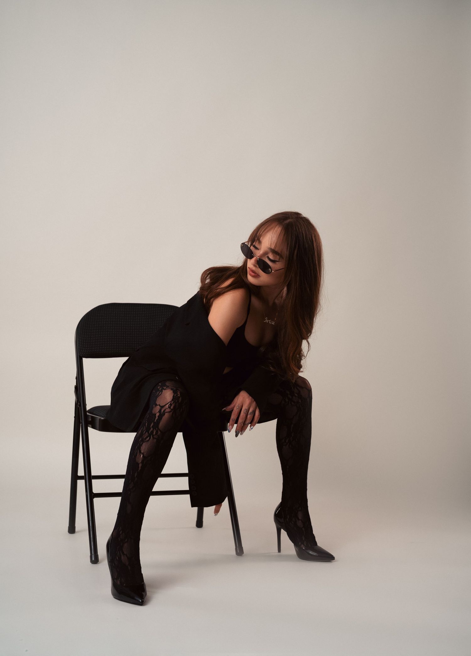 Model wearing black boots and leggings poses artistically with chair against white backdrop.