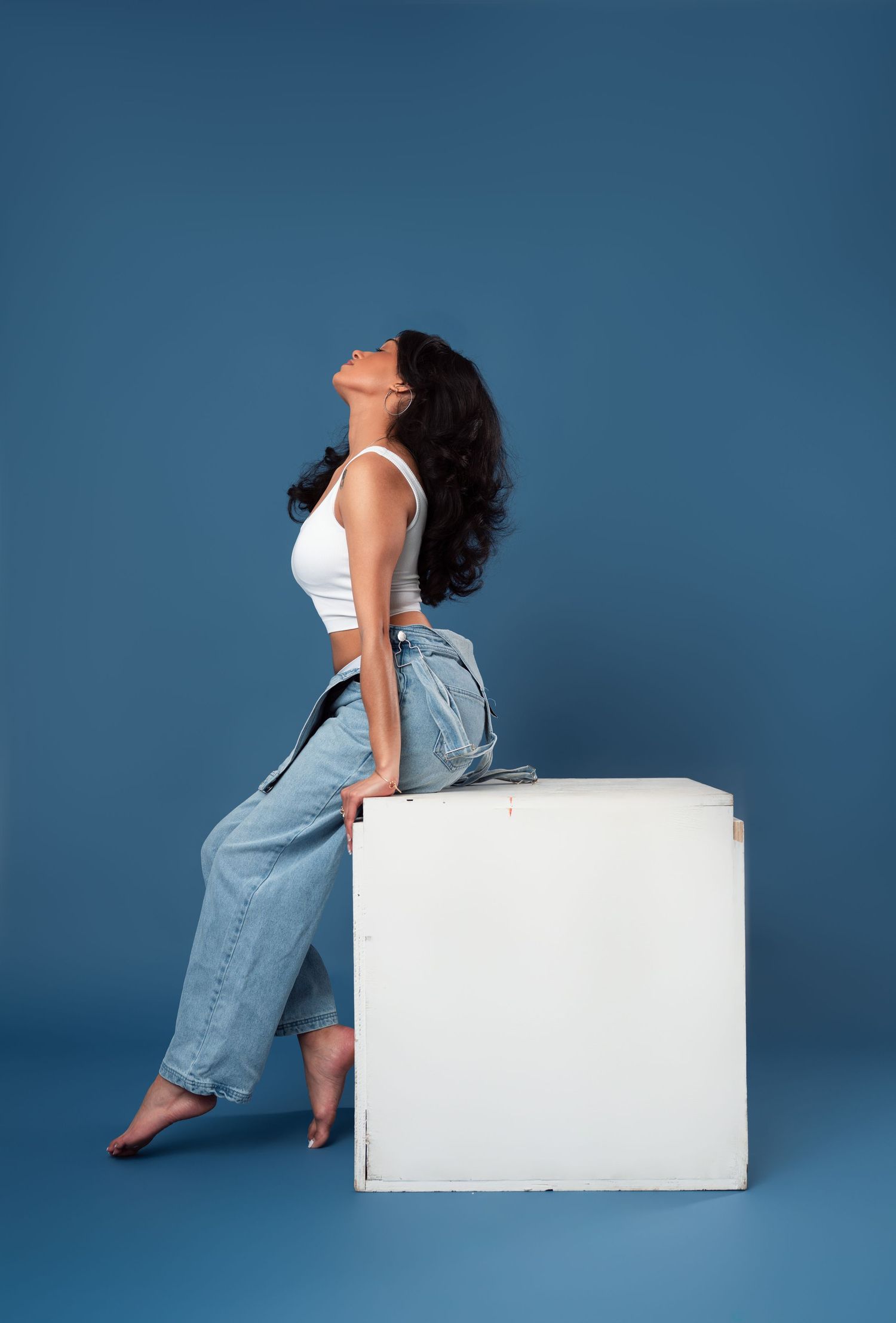 Someone in casual white top and light blue jeans poses sitting on a white cube against a dark blue studio background.