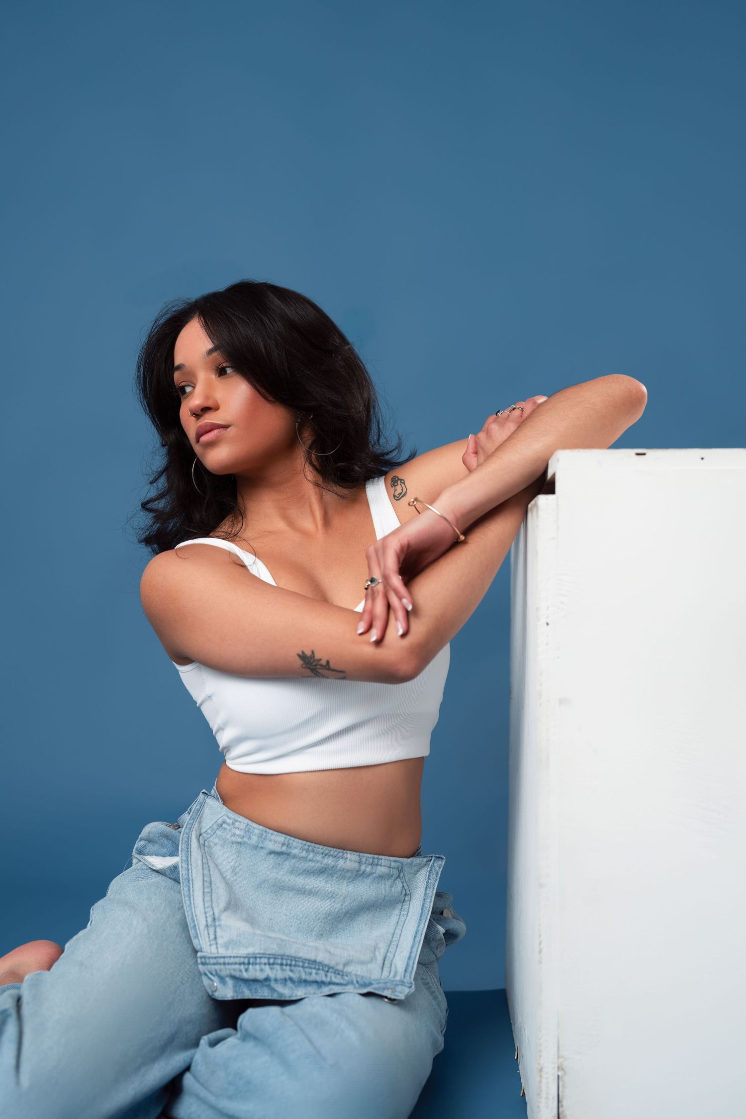 Fashion model in white crop top and light denim poses against blue studio backdrop.