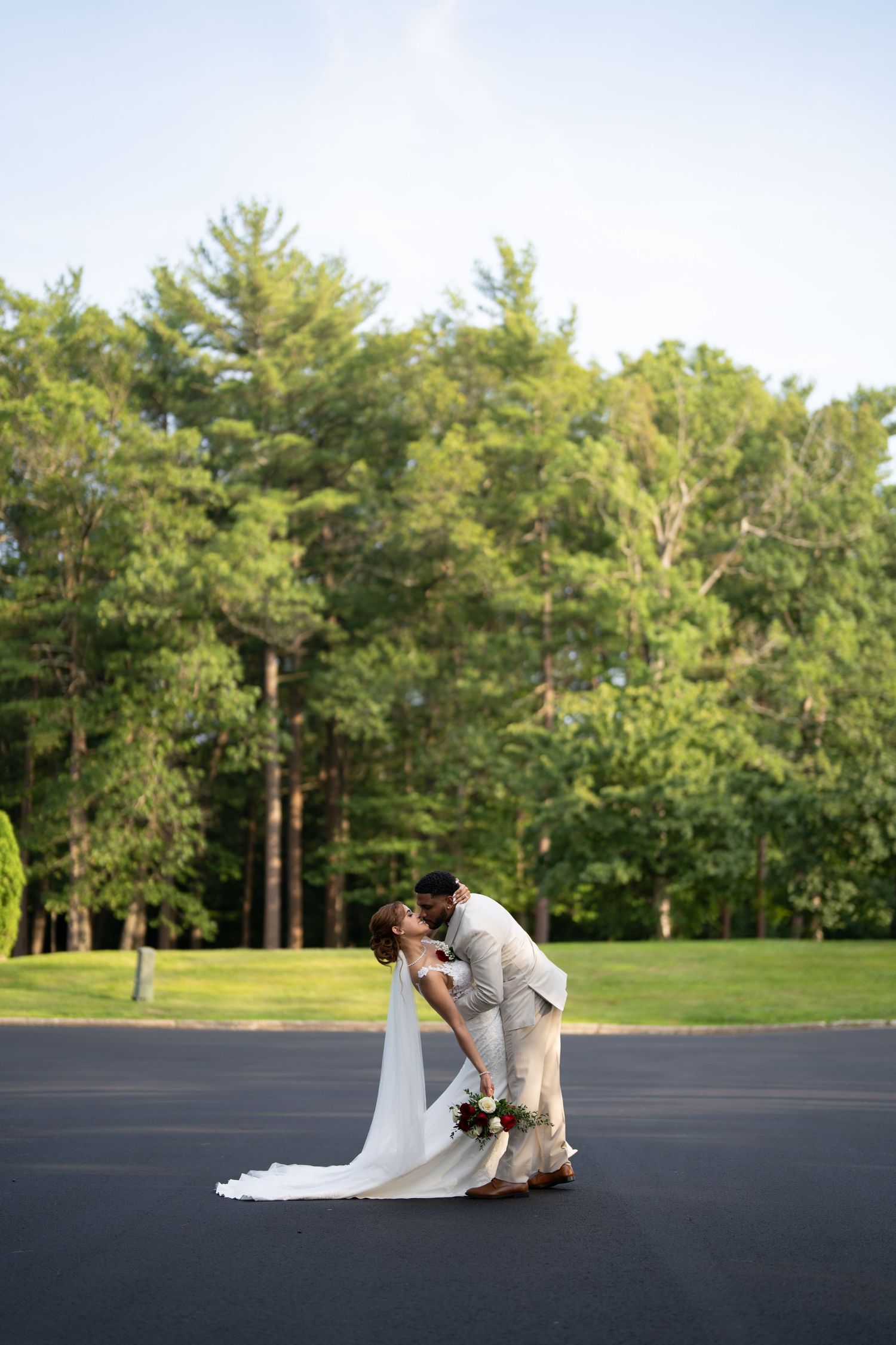 A romantic dance dip sequence between a couple in wedding attire on a blacktop surrounded by pine trees.