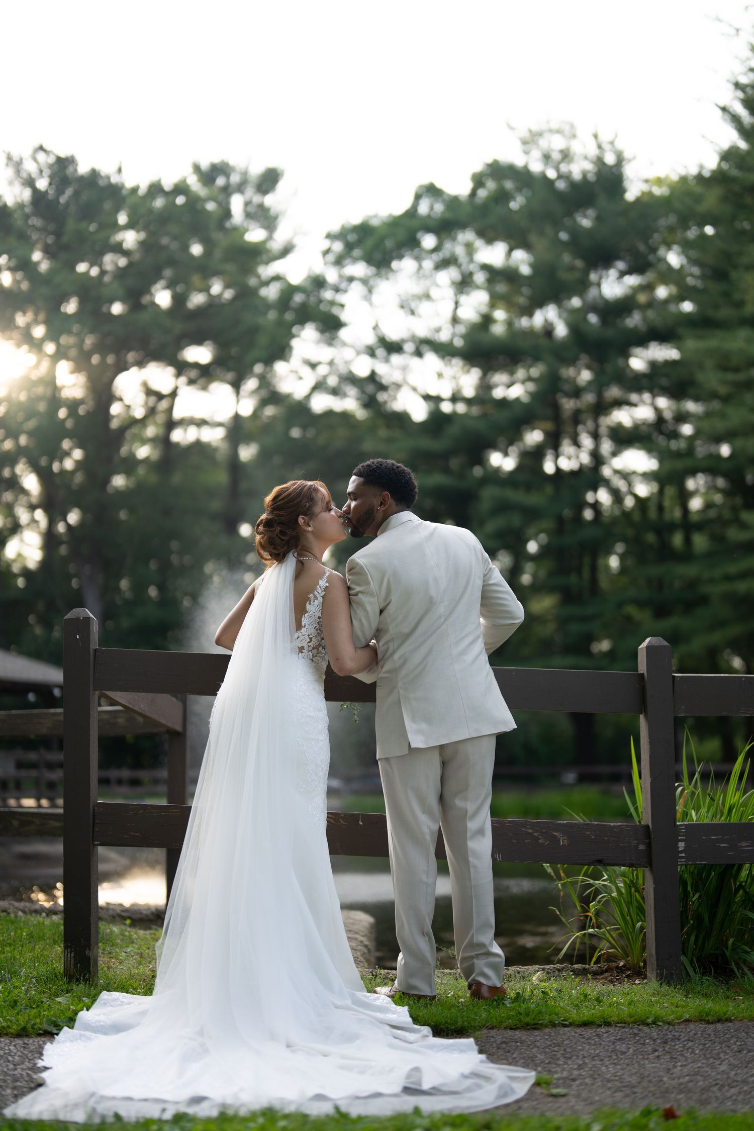 A bride and groom share a romantic moment at sunset by a wooden fence with pine trees in the background.