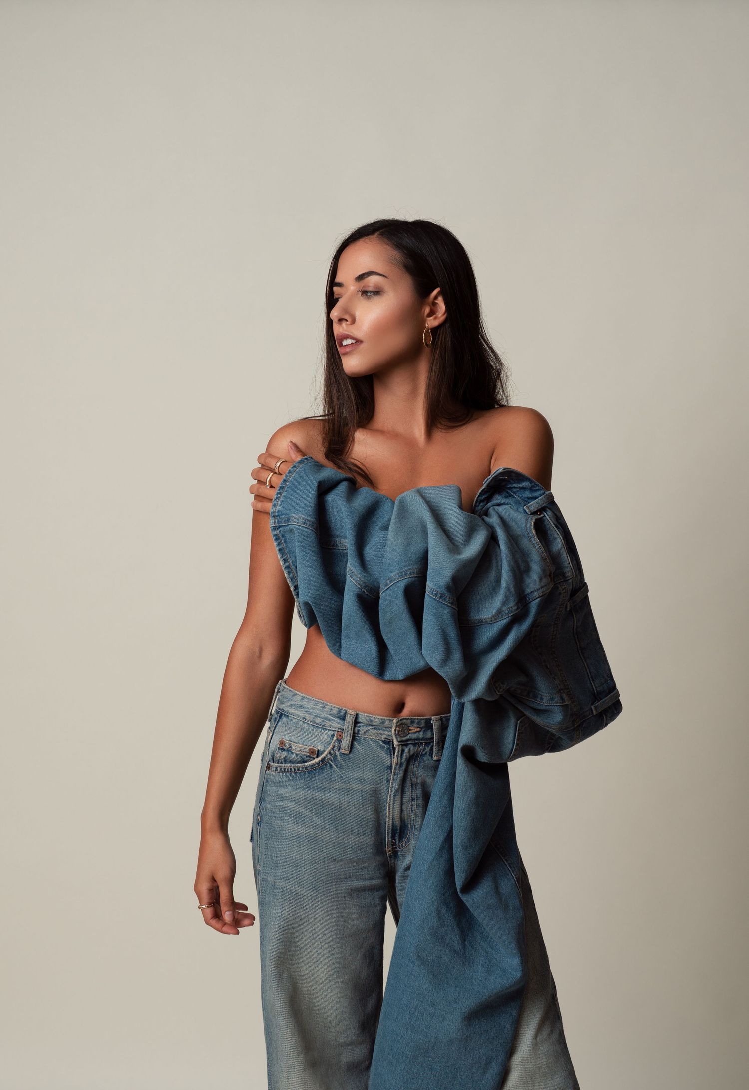 Fashion model poses in denim outfit featuring ruffled asymmetrical top and high-waisted jeans against neutral backdrop.