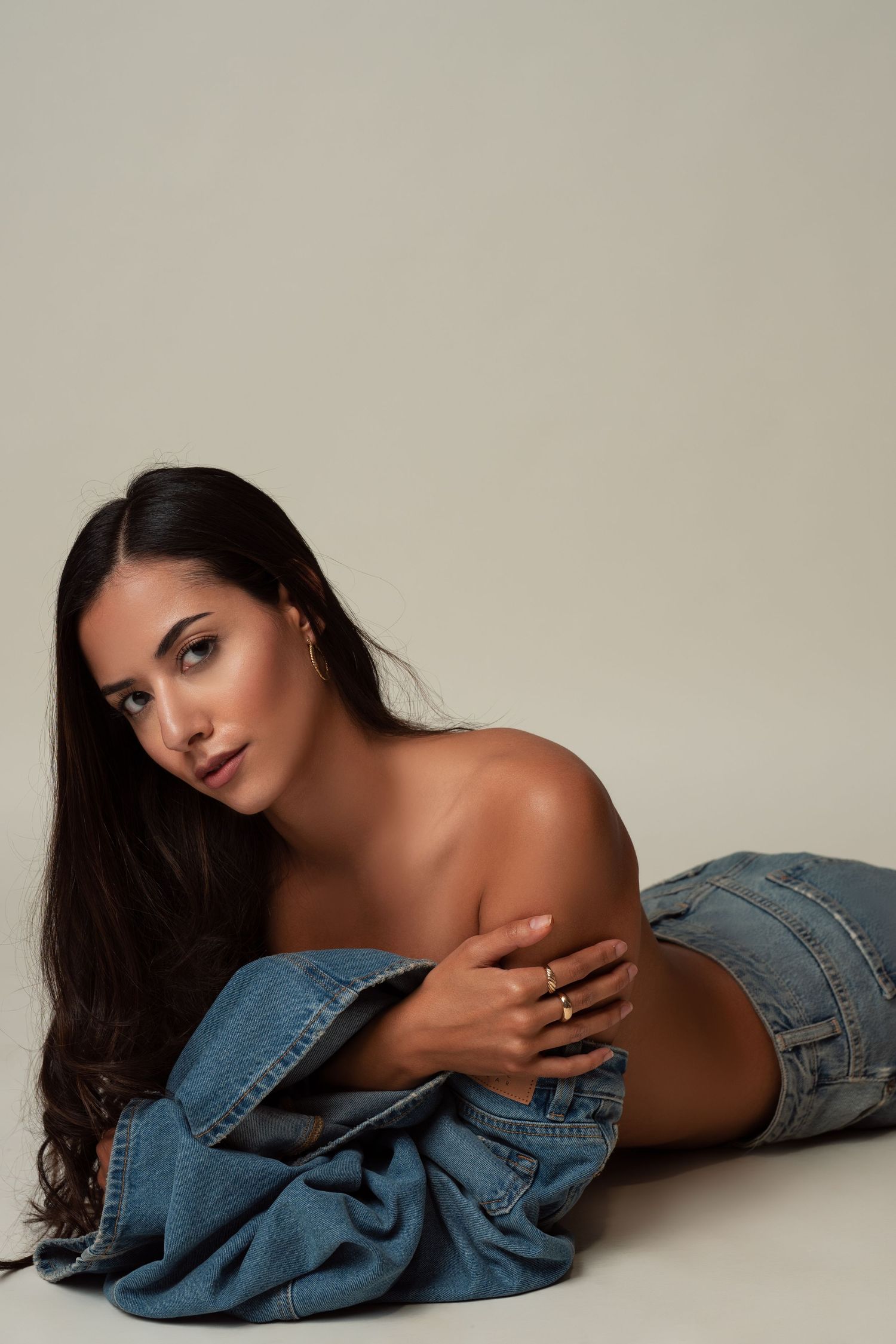 Woman with long dark hair poses artistically wearing denim jacket against neutral background in fashion photo series.