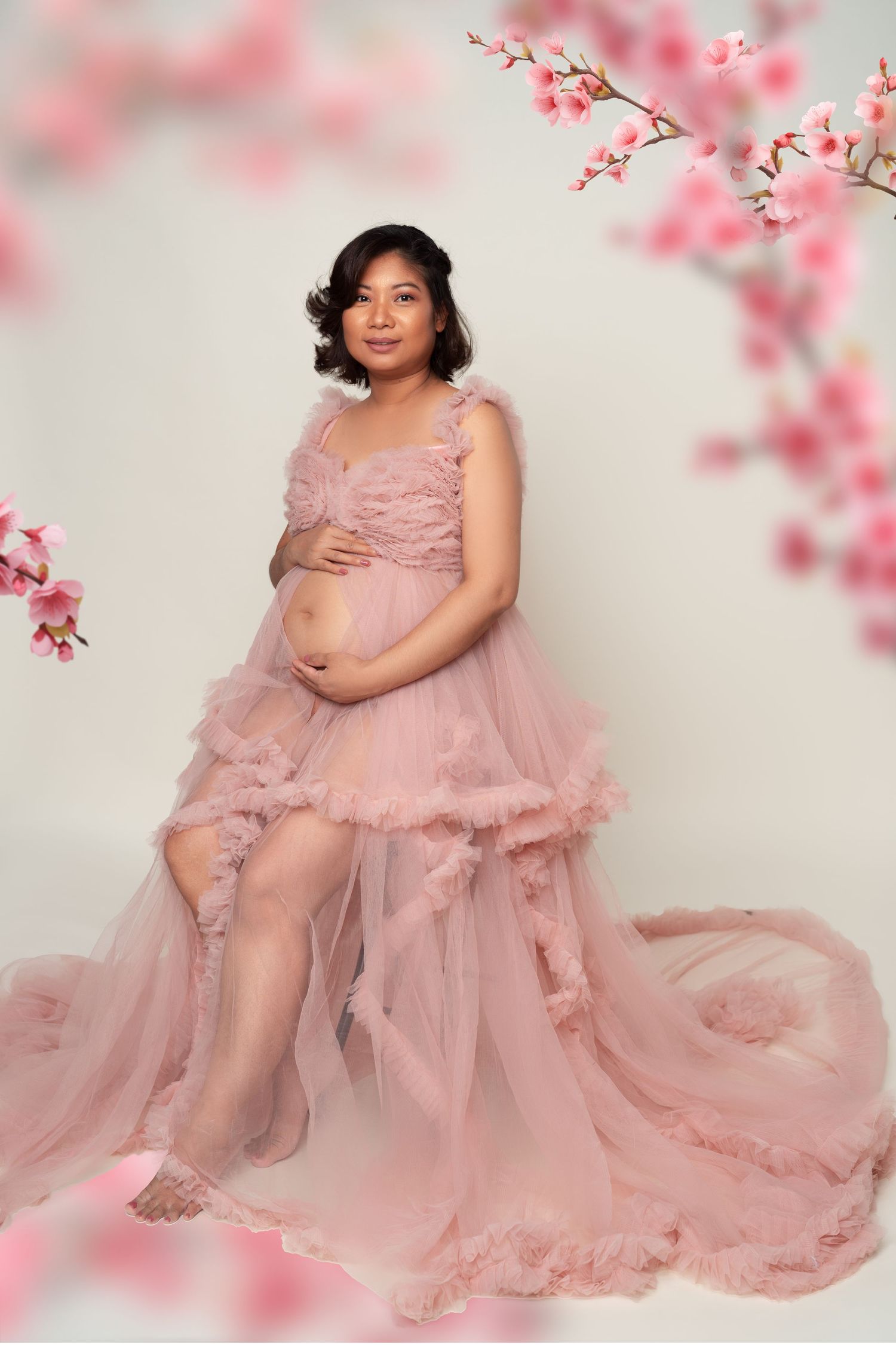 A maternity portrait with flowing pink tulle gown and cherry blossoms framing the dreamy scene.
