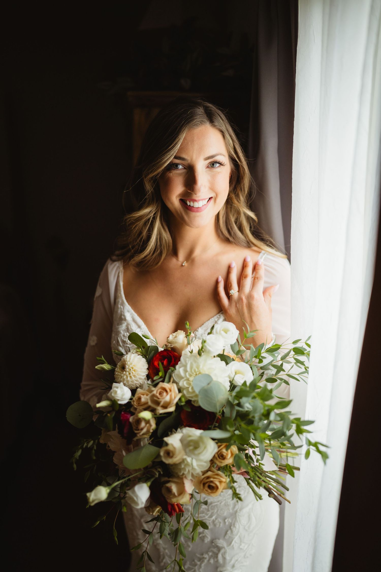 A wedding bouquet featuring white peonies, roses and greenery is held near a window with soft natural light.