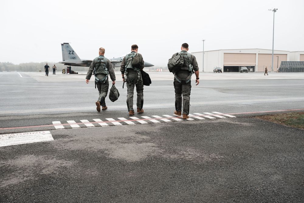 Military personnel walking across wet tarmac with aircraft visible in background.