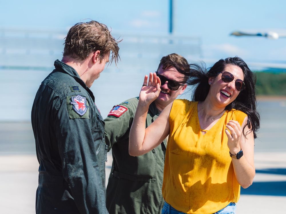Group celebrating with pilot in flight suit on sunny airfield tarmac.