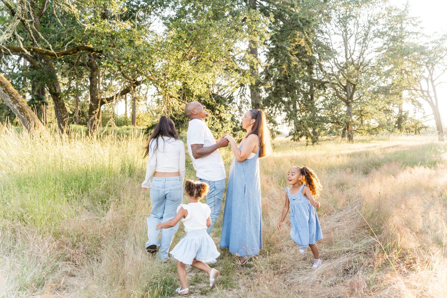 A family in coordinating blue and white outfits walk together through a sunlit meadow on a summer evening during a family photo session.