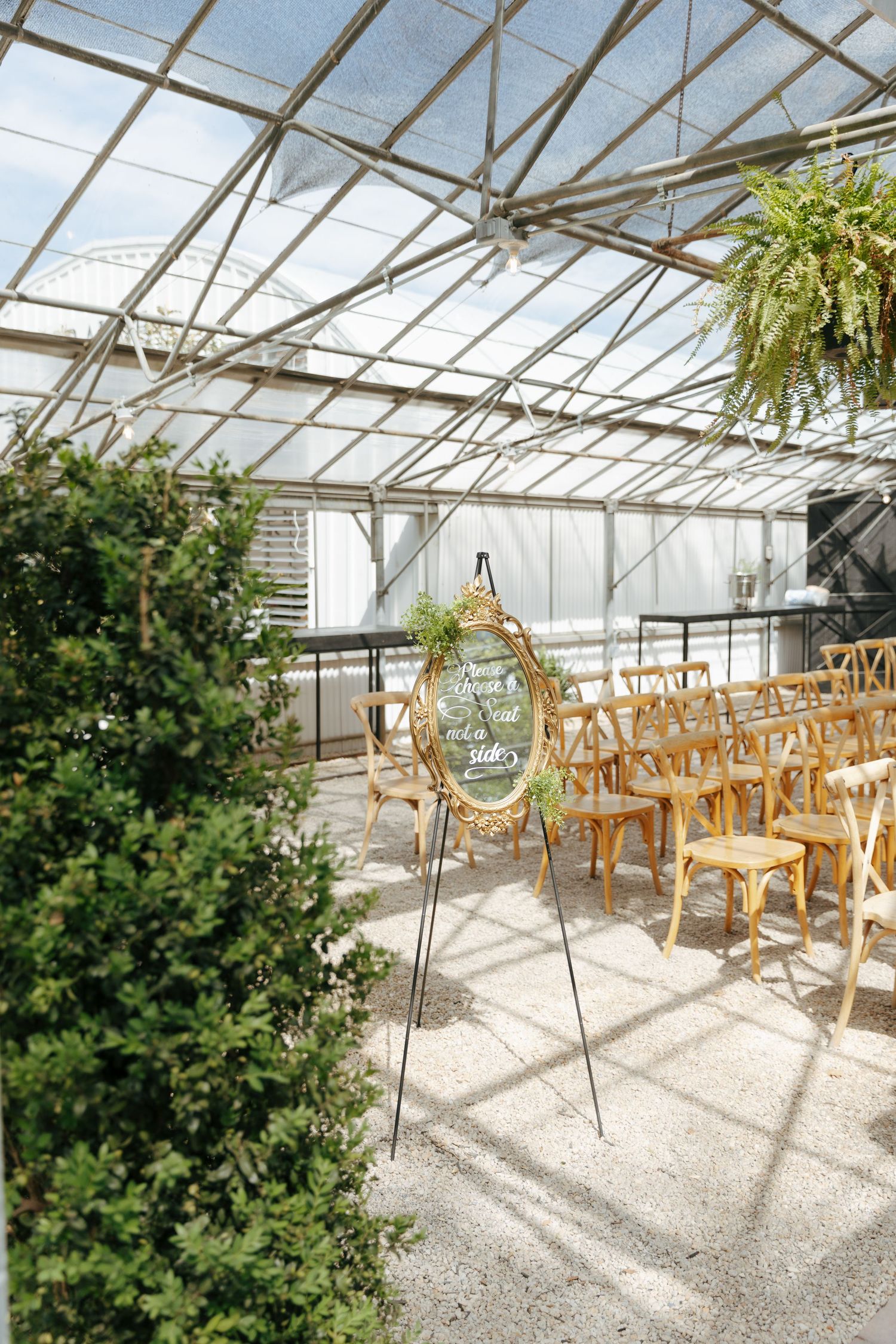 Glass-enclosed greenhouse interior with wooden chairs arranged on gravel floor and greenery visible along the sides.