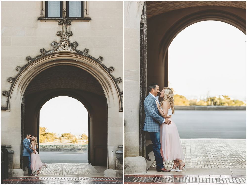 A couple shares a romantic moment under an arched passageway overlooking a coastal view at sunset.