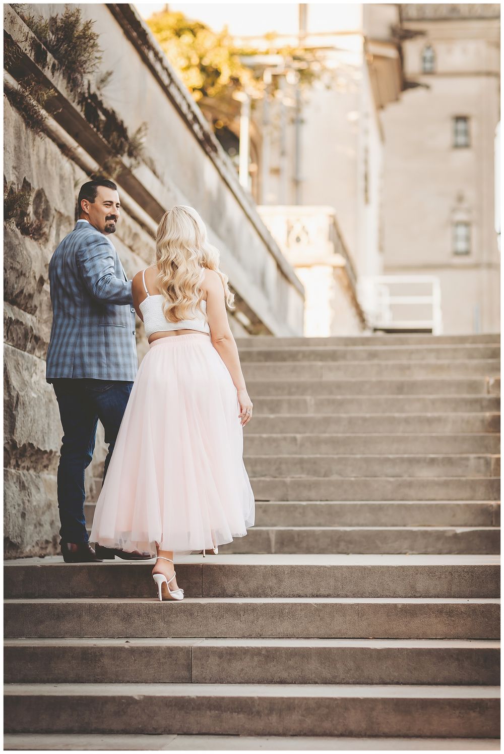 A couple stands on stone steps, with the bride wearing a pink tulle skirt and the groom in a plaid blazer.