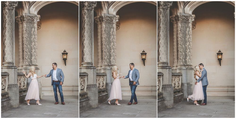 Romantic couple in elegant attire share tender moments in front of an ornate stone archway at a historic venue.