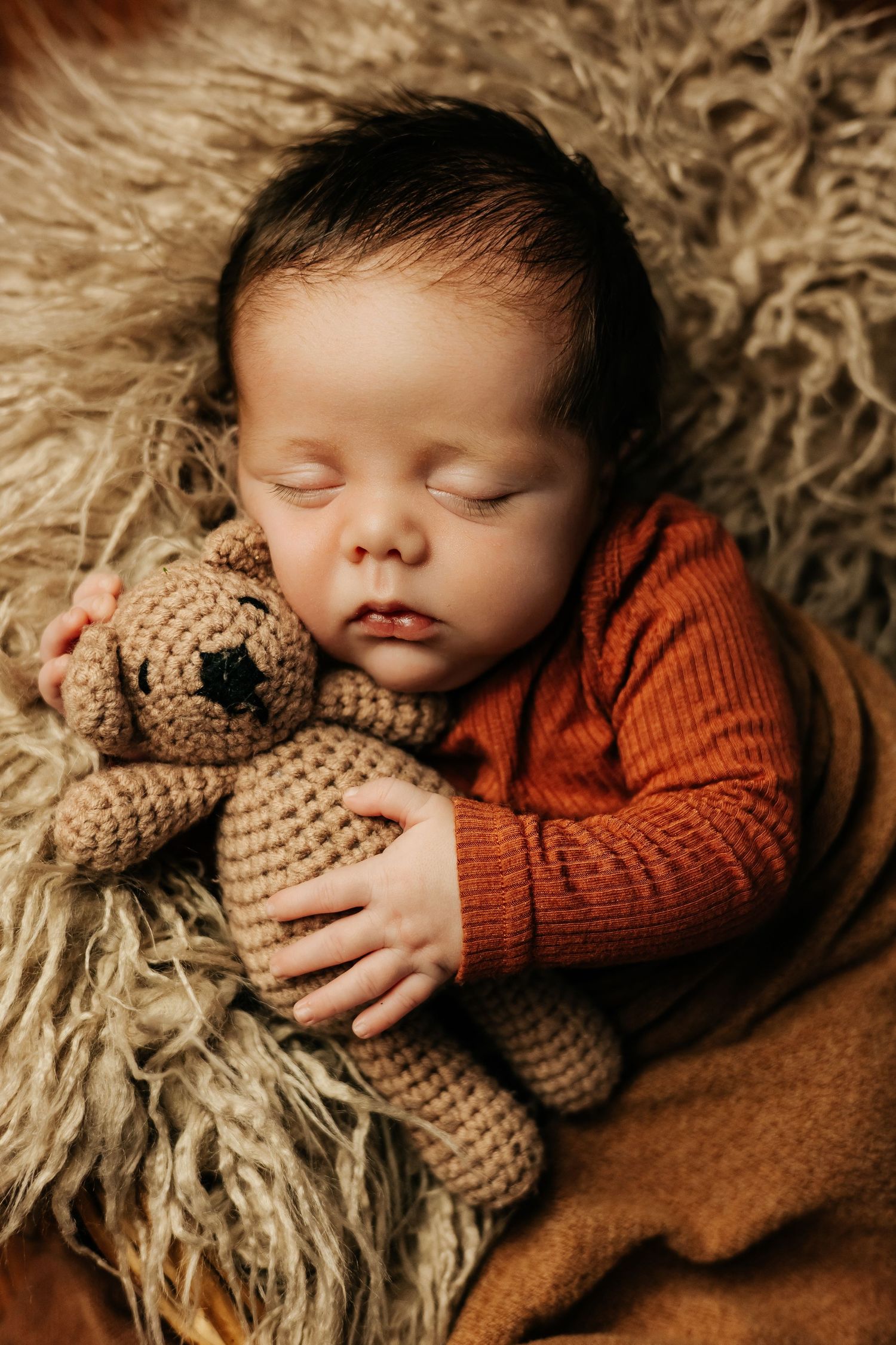 Sleeping newborn in orange sweater cuddling brown teddy bear on furry backdrop.