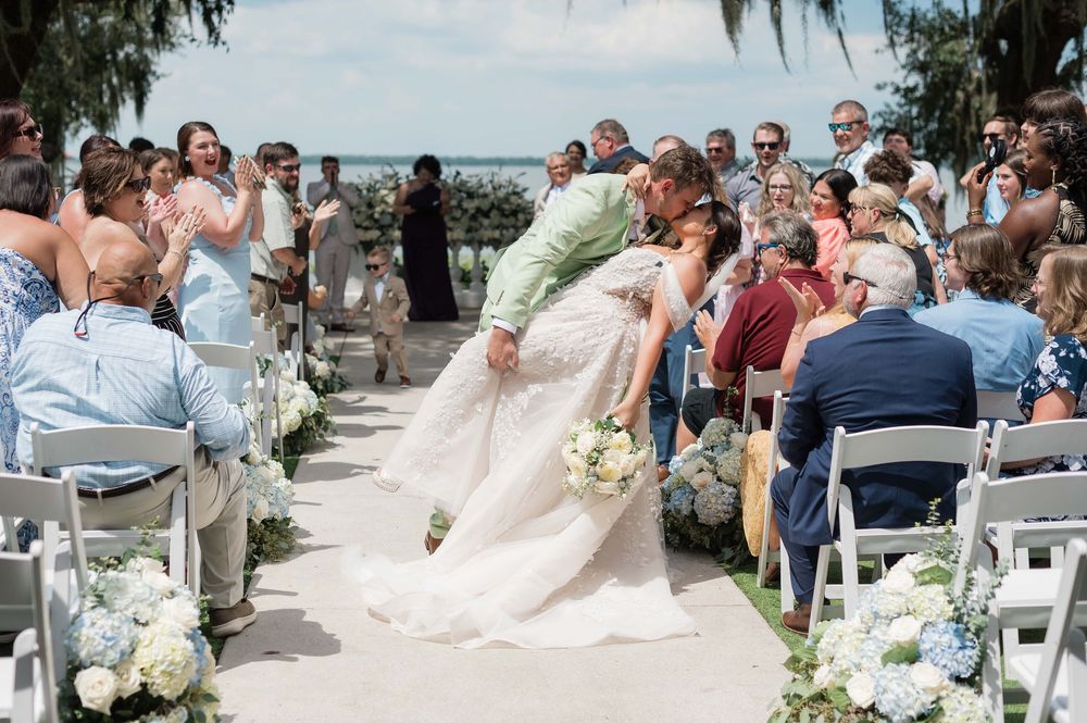 Bride and groom share a romantic dip kiss at outdoor beach wedding ceremony with guests applauding.