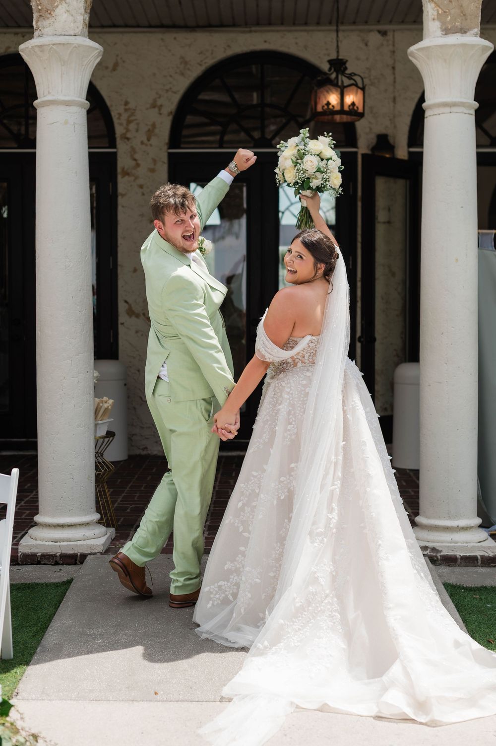 Joyful newlyweds celebrate after wedding ceremony, bride in lace gown holding bouquet, groom in mint green suit.
