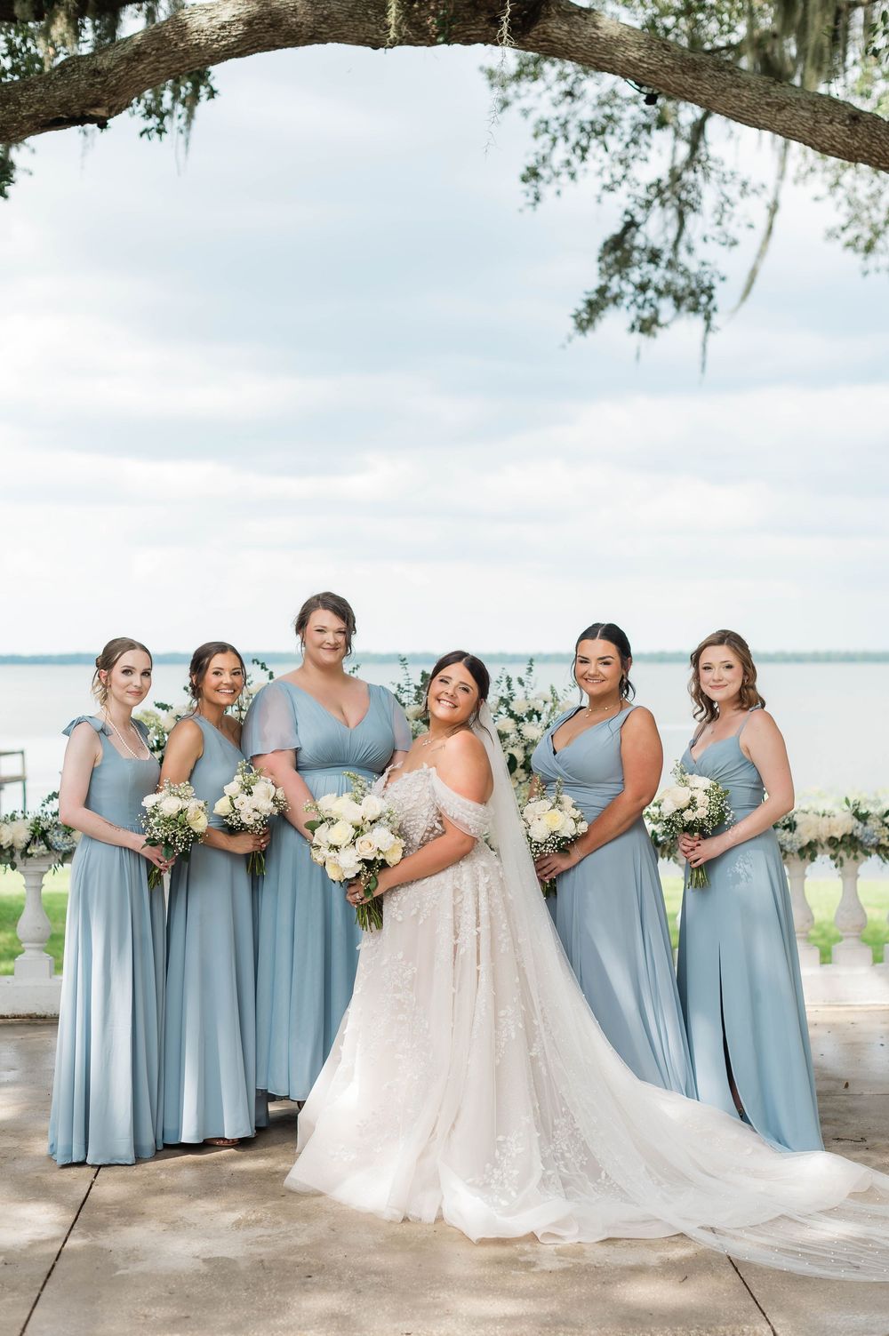 Bride in lace gown with five bridesmaids in dusty blue dresses holding white bouquets outdoors.