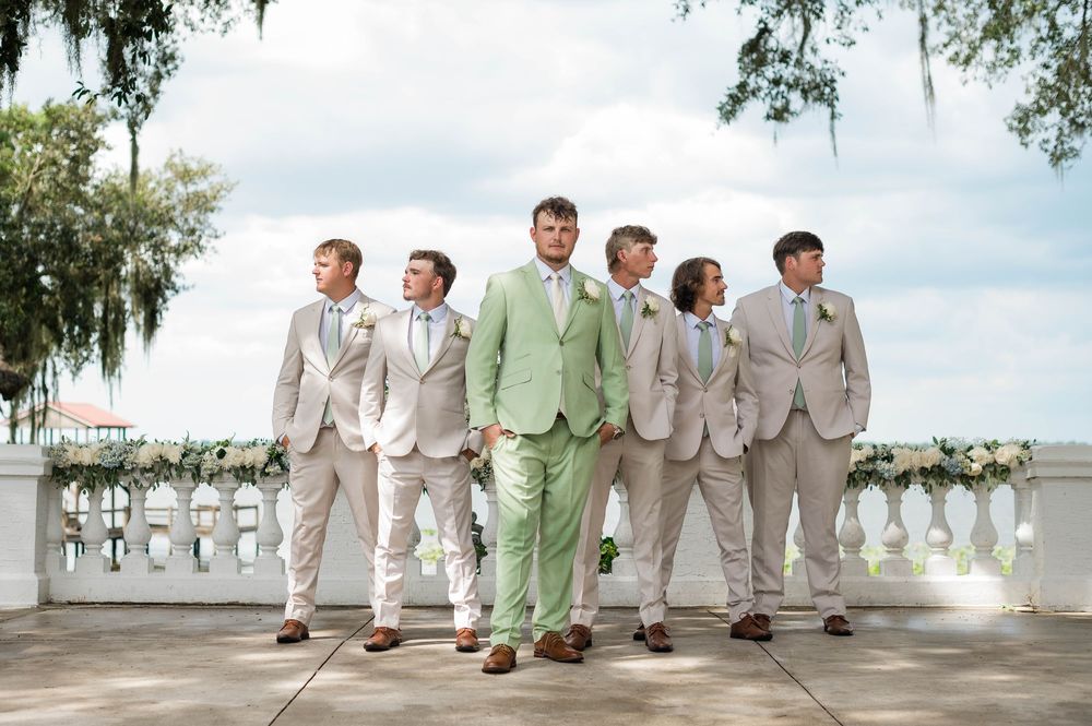 Groom in mint green suit poses with groomsmen in tan suits on elegant outdoor venue balcony.