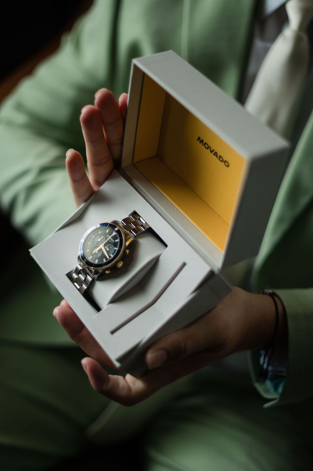 Groom in mint green suit holding open a Movado watch box displaying a silver and green chronograph watch.