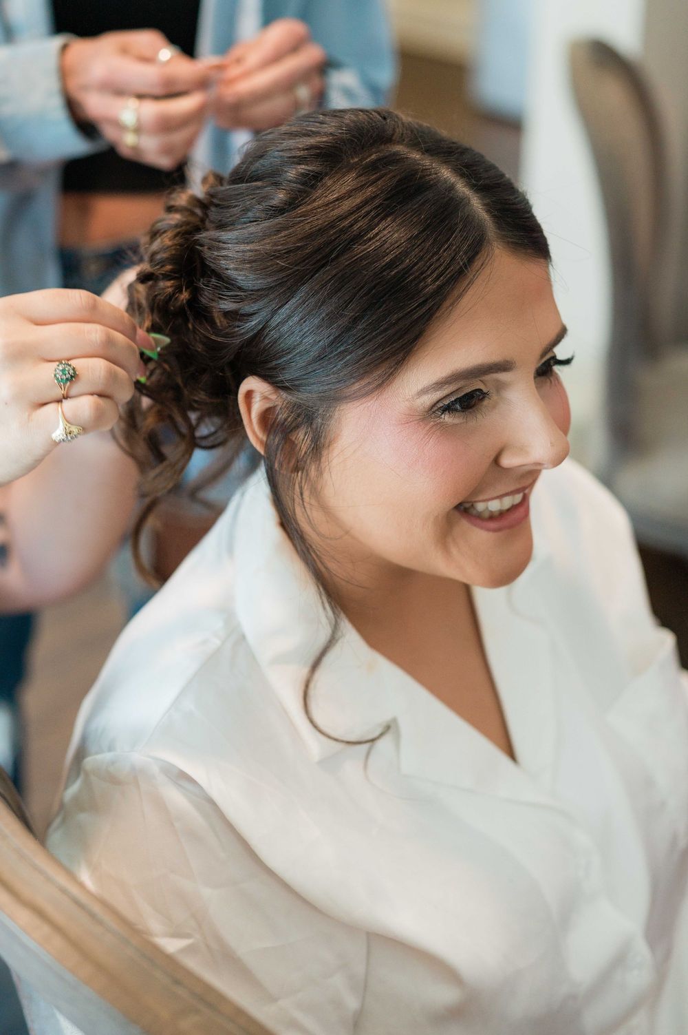 Bride smiling during hair styling session in white robe on wedding day.