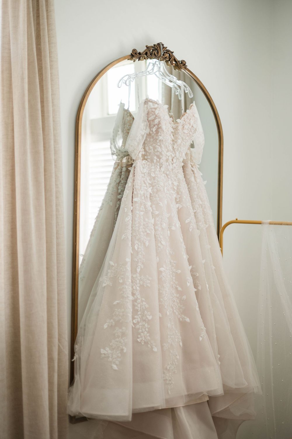 Blush lace wedding gown hanging on an ornate gold arched mirror with sheer curtains in a bright bridal suite.