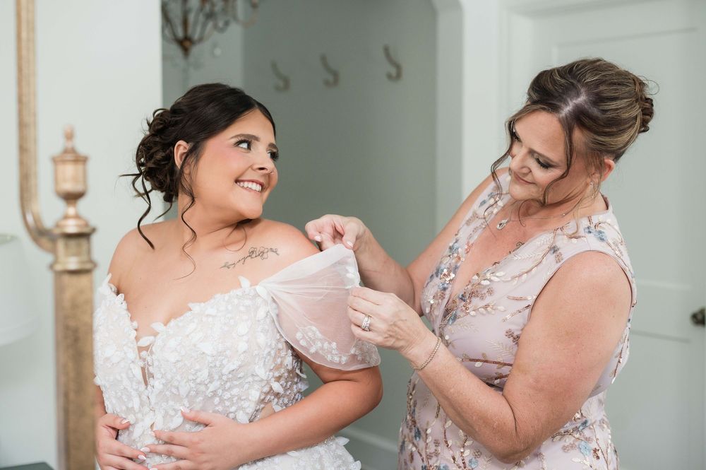 Bride in lace wedding dress gets help adjusting gown from woman in floral dress during wedding preparation.