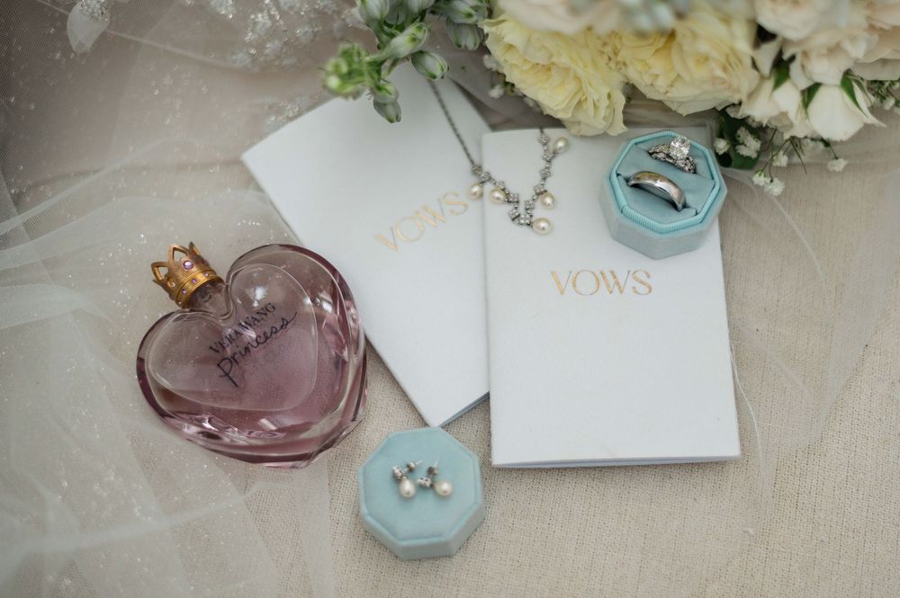 Wedding flat lay with vows books, diamond rings, pearl earrings, silver necklace, and pink perfume bottle beside white flowers.
