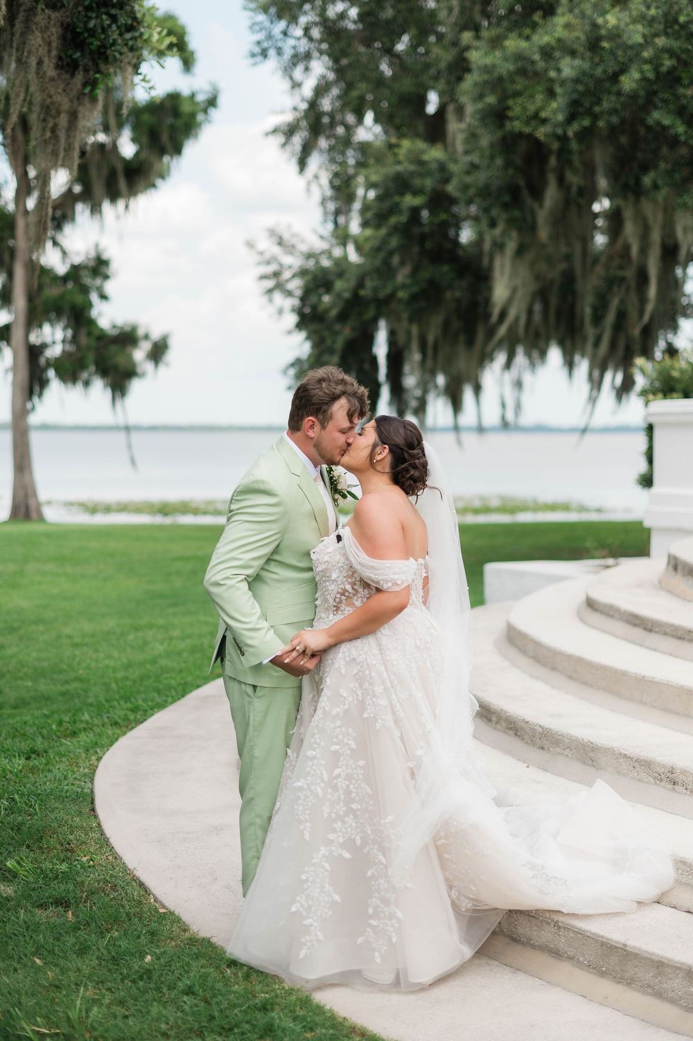 Bride and groom sharing kiss in lush garden by waterfront in elegant wedding attire.
