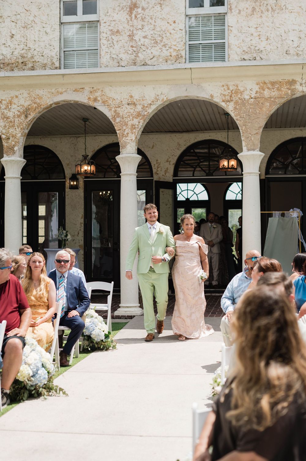 Groom in mint suit walks down outdoor wedding aisle with mother at elegant venue with arched colonnade.