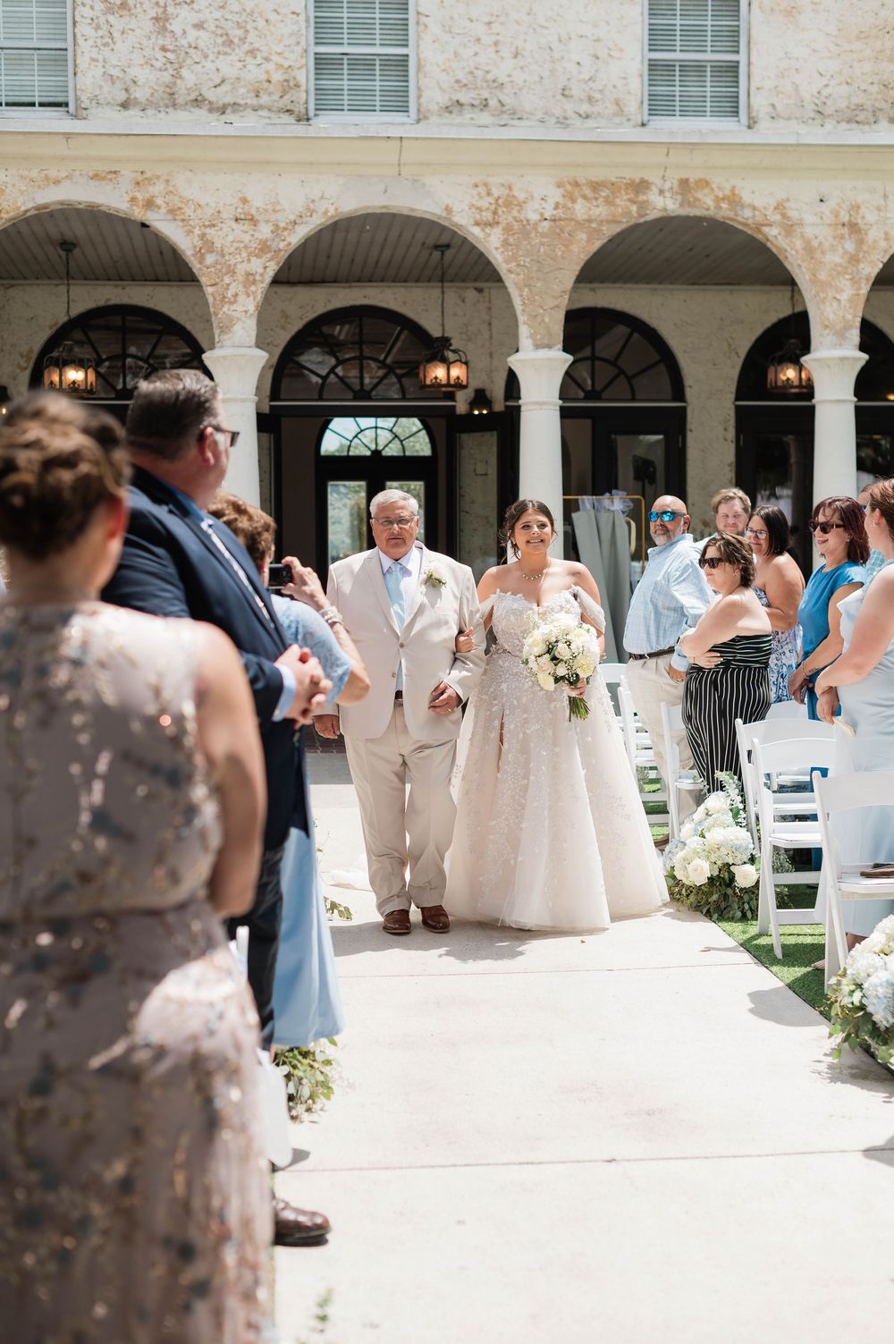 Bride walks down outdoor aisle with father during sunny wedding ceremony at elegant venue with arched colonnade.