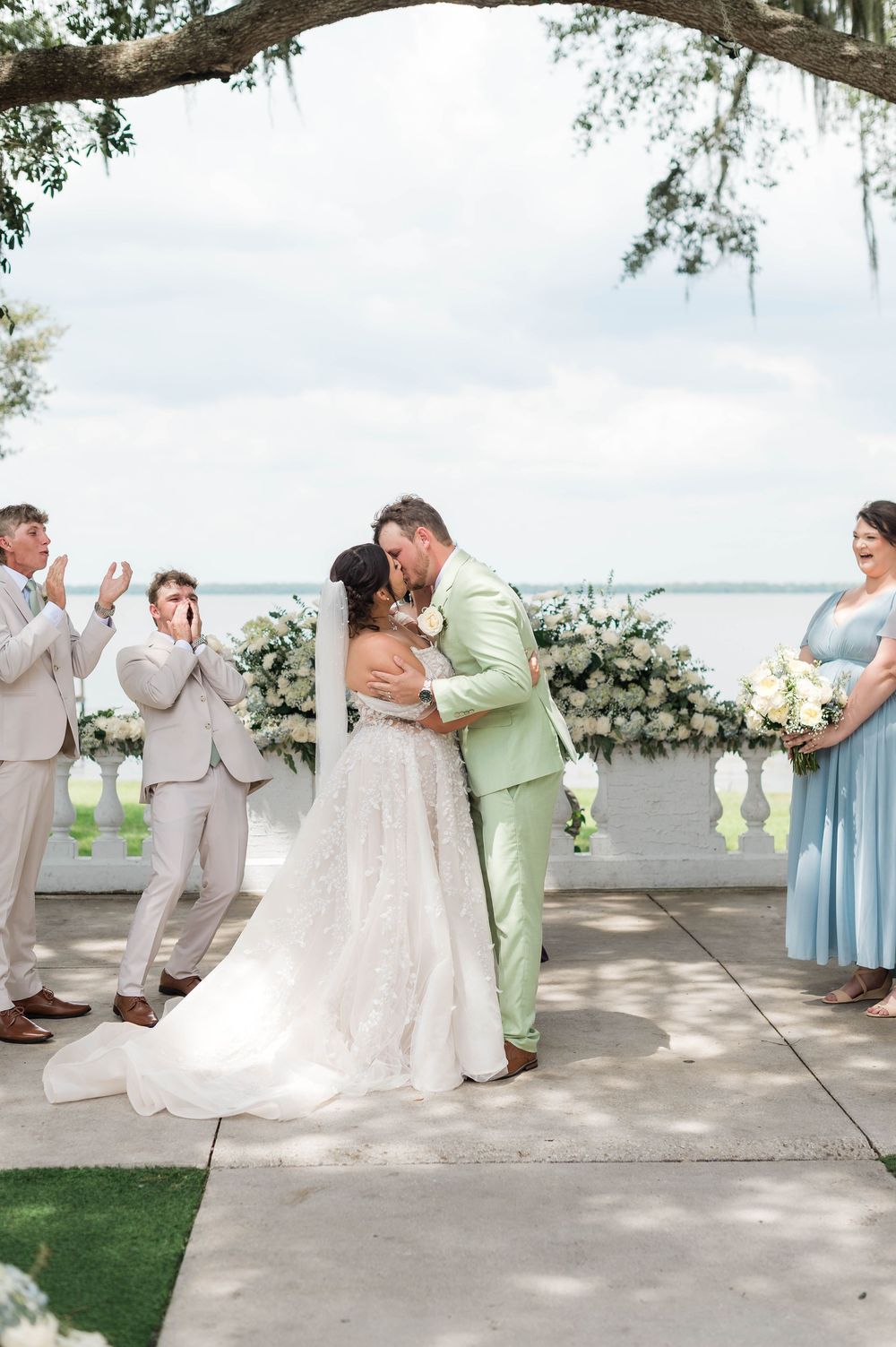 Bride and groom share first kiss at outdoor waterfront ceremony with wedding party in sage and blue attire.
