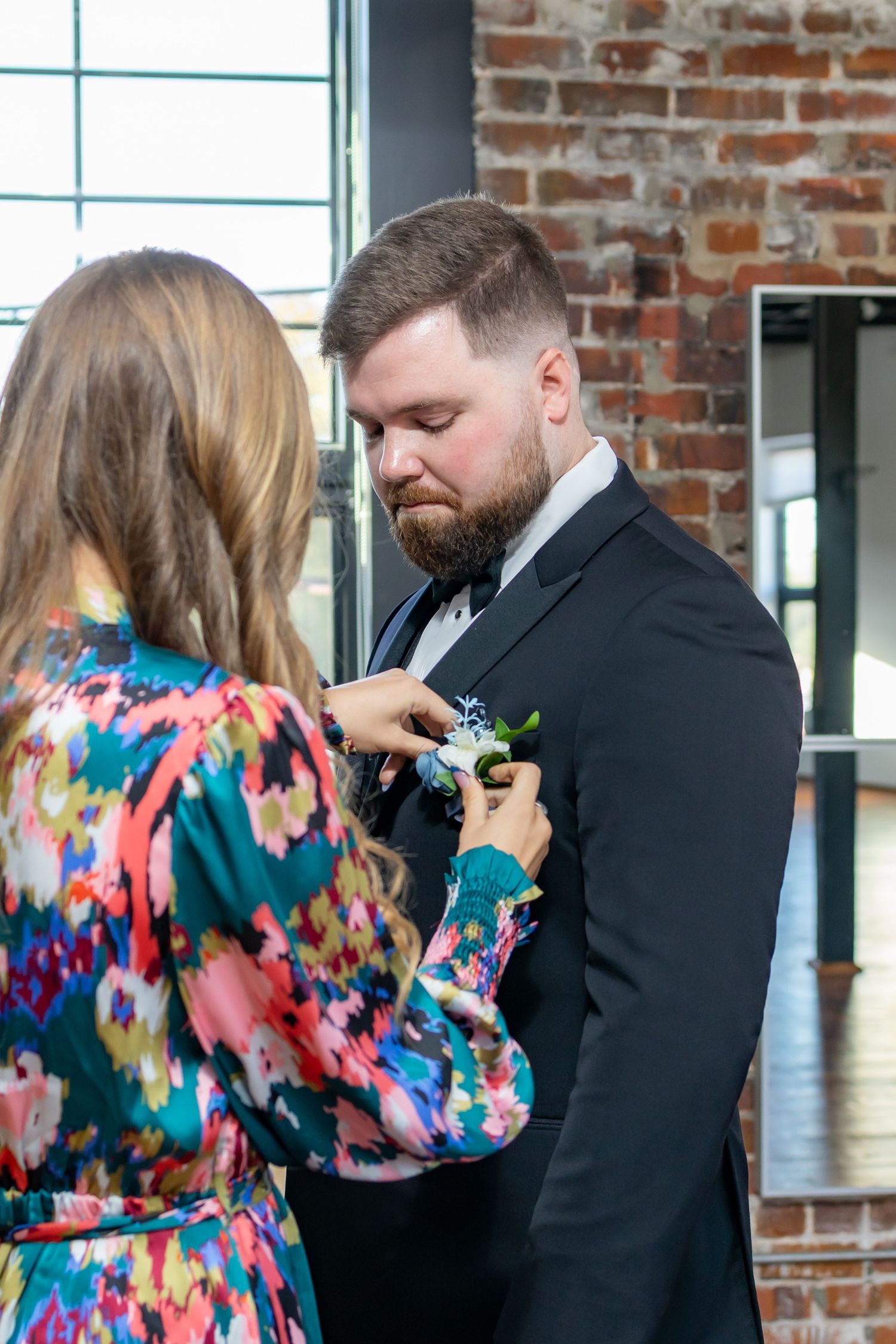 A couple in formal attire shares a first look moment against a brick wall backdrop in a sunlit room.