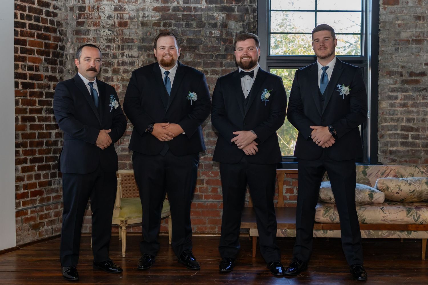 Groomsmen in dark suits standing together in front of exposed brick wall at wedding venue.