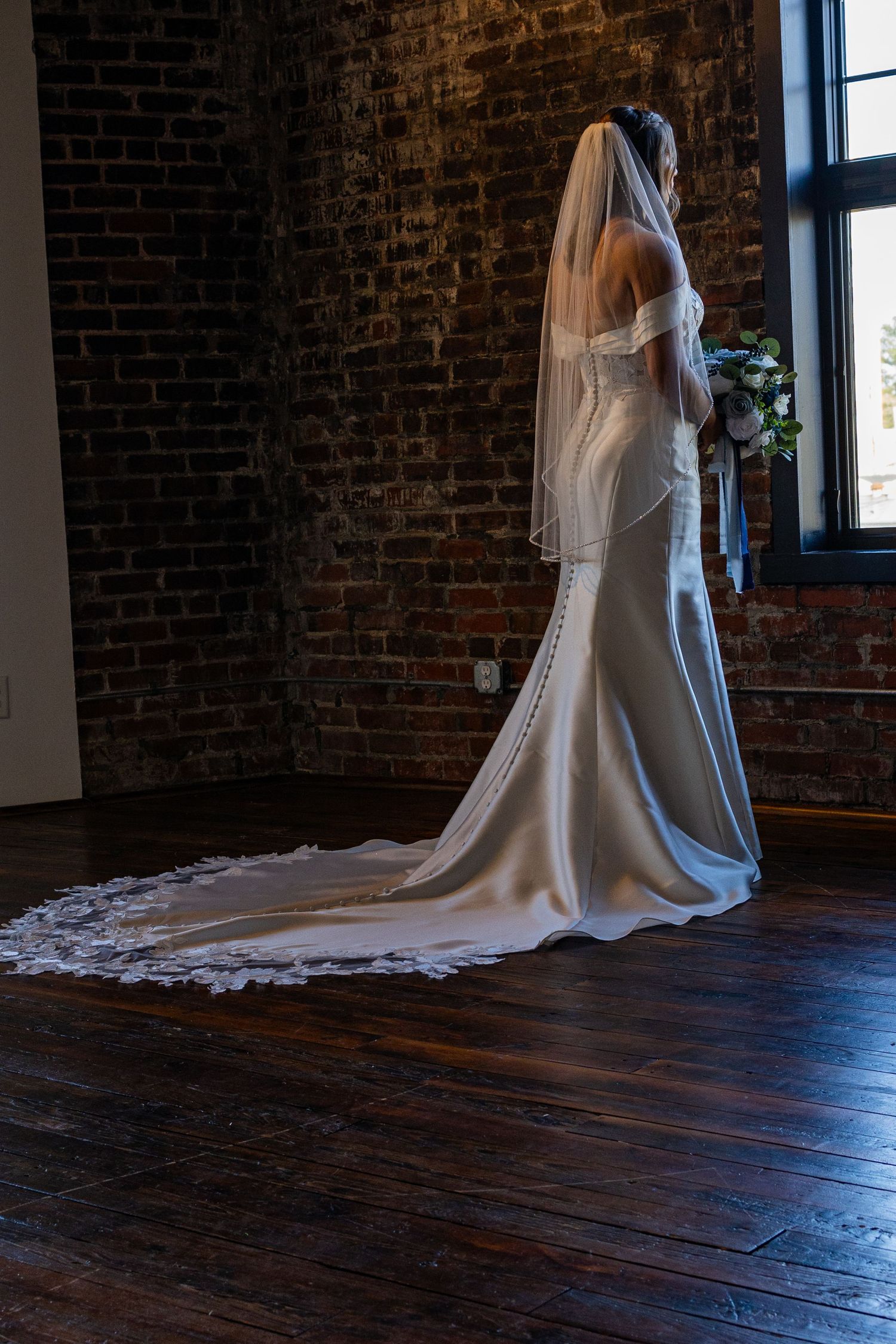 A bride in a white wedding dress with cathedral-length veil stands gracefully against exposed brick walls and hardwood floors.