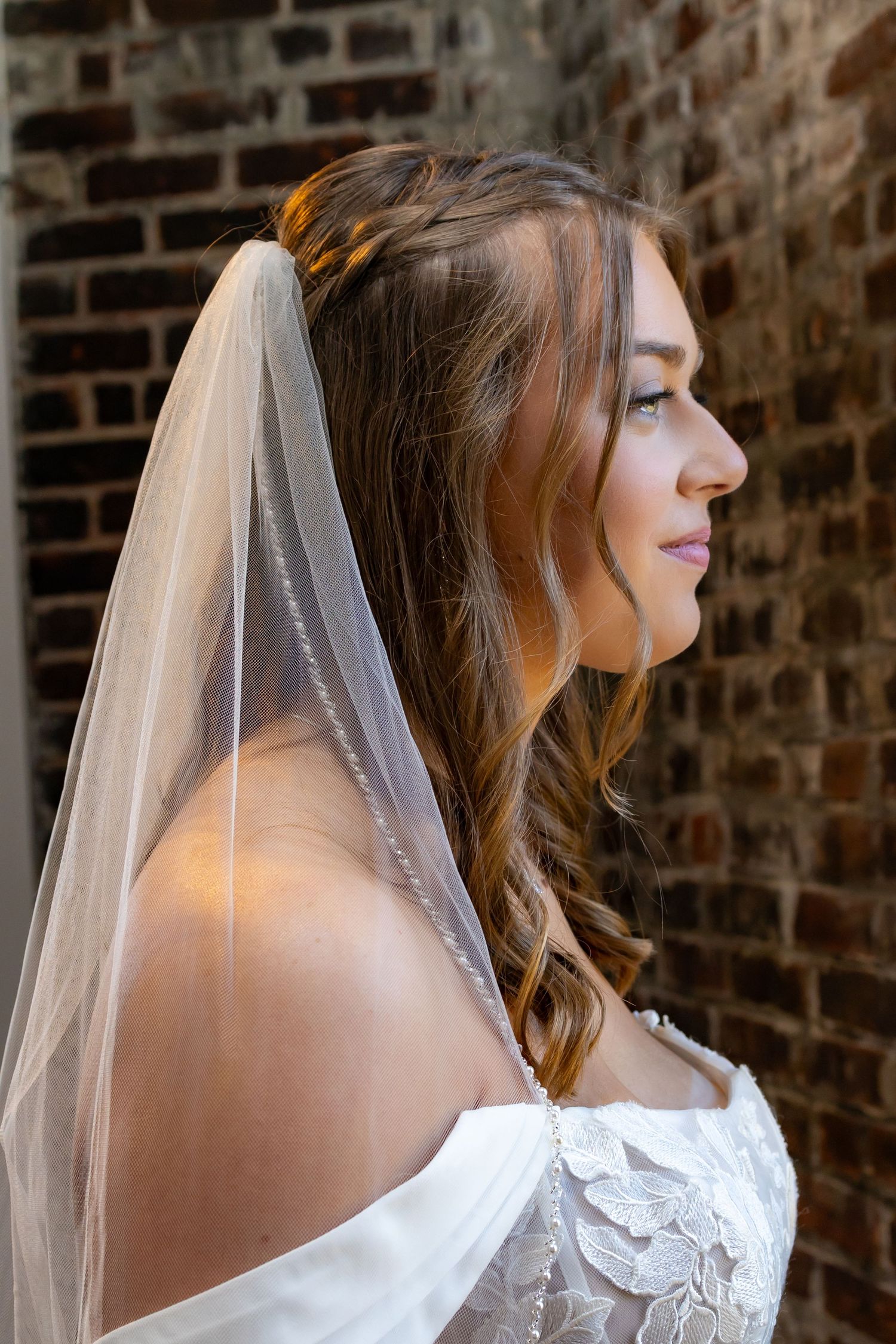 Elegant bridal portrait against exposed brick wall showcases delicate lace details and flowing veil in natural light.