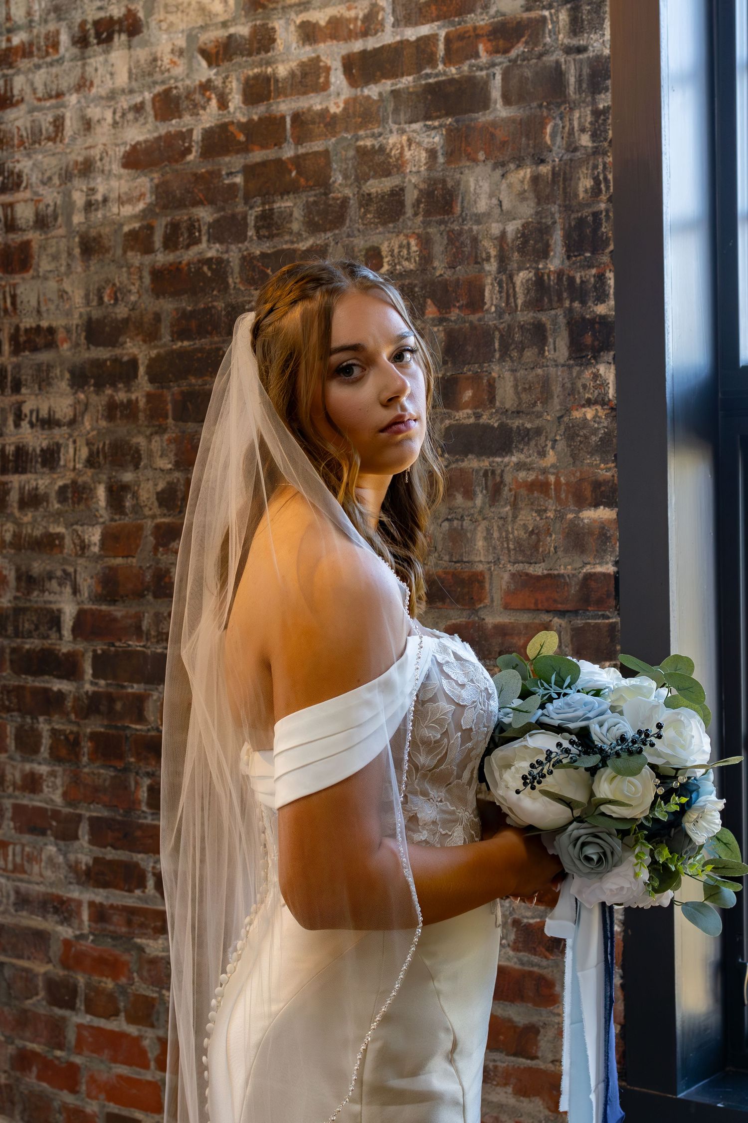 Bride posing in an off-shoulder white wedding gown against exposed brick walls holding a white and green bouquet.