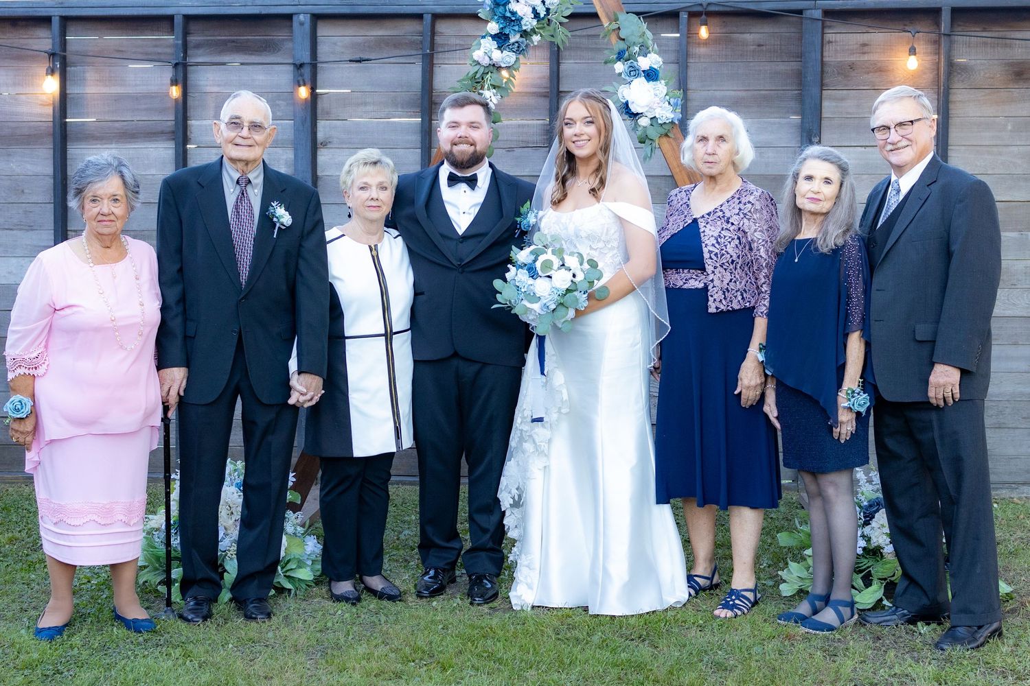 A large outdoor family wedding portrait with guests lined up in formal attire against a wooden wall backdrop.