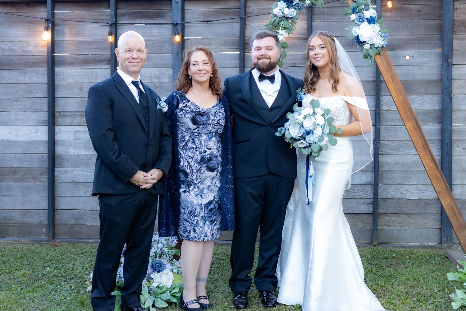 Wedding party poses outdoors with bride in white dress and blue floral bouquet against wooden wall.