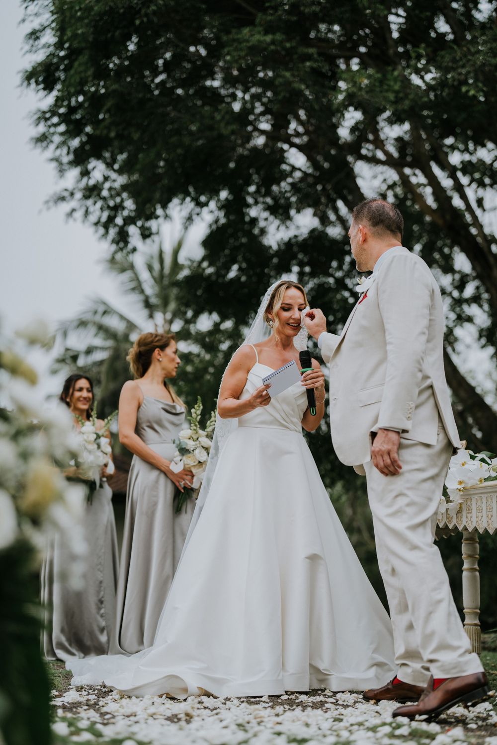 Wedding party members in elegant white dresses and suits gather on a flower-strewn path.