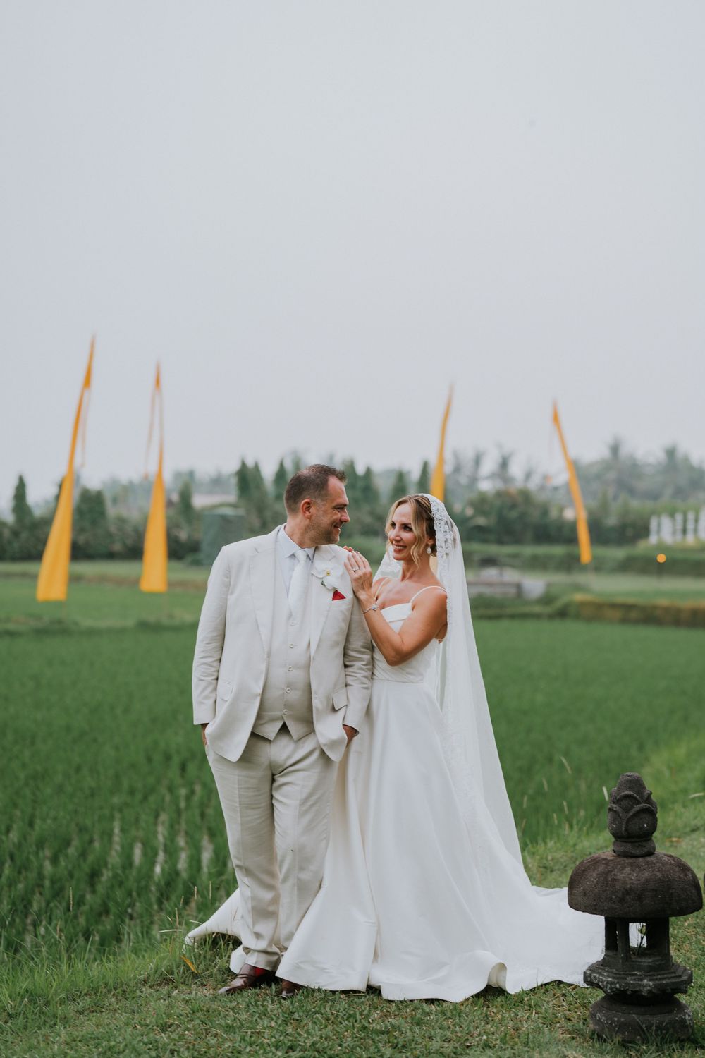 A wedding couple poses in a lush green rice field with tall yellow banners and a stone lantern in Bali.