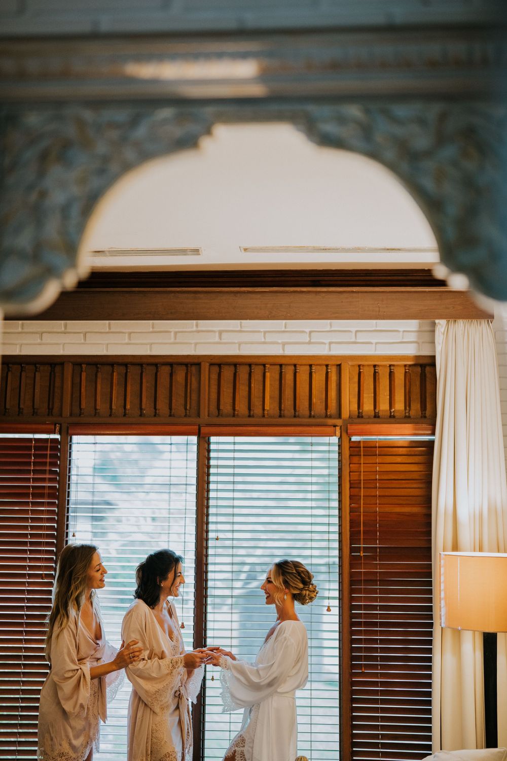 Three women in robes stand by a window during wedding preparations under an ornate archway.