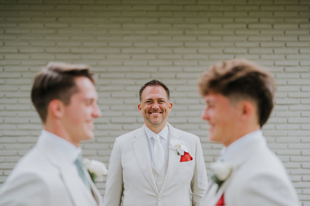 Three men in light colored suits share a moment against a white brick wall.
