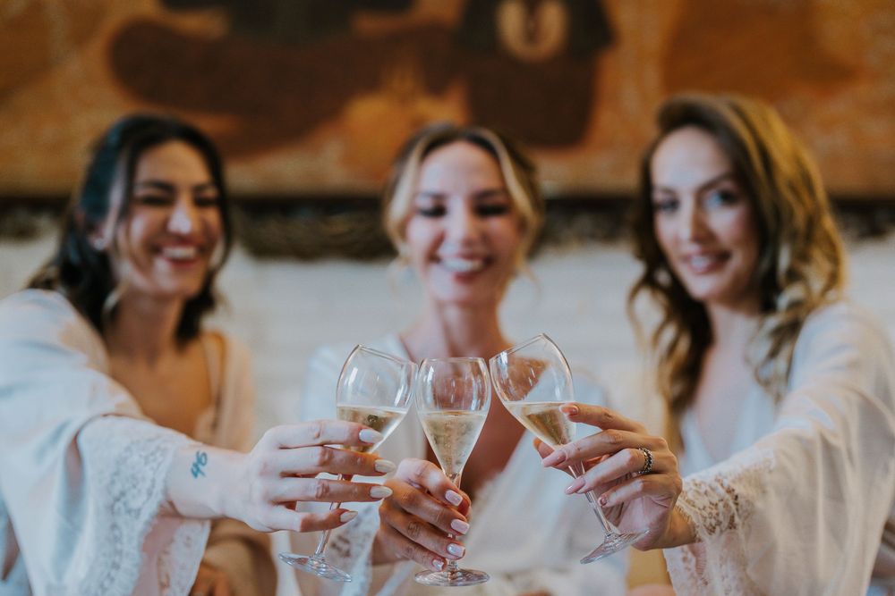 Three women in white robes toast with wine glasses during pre-wedding celebrations.