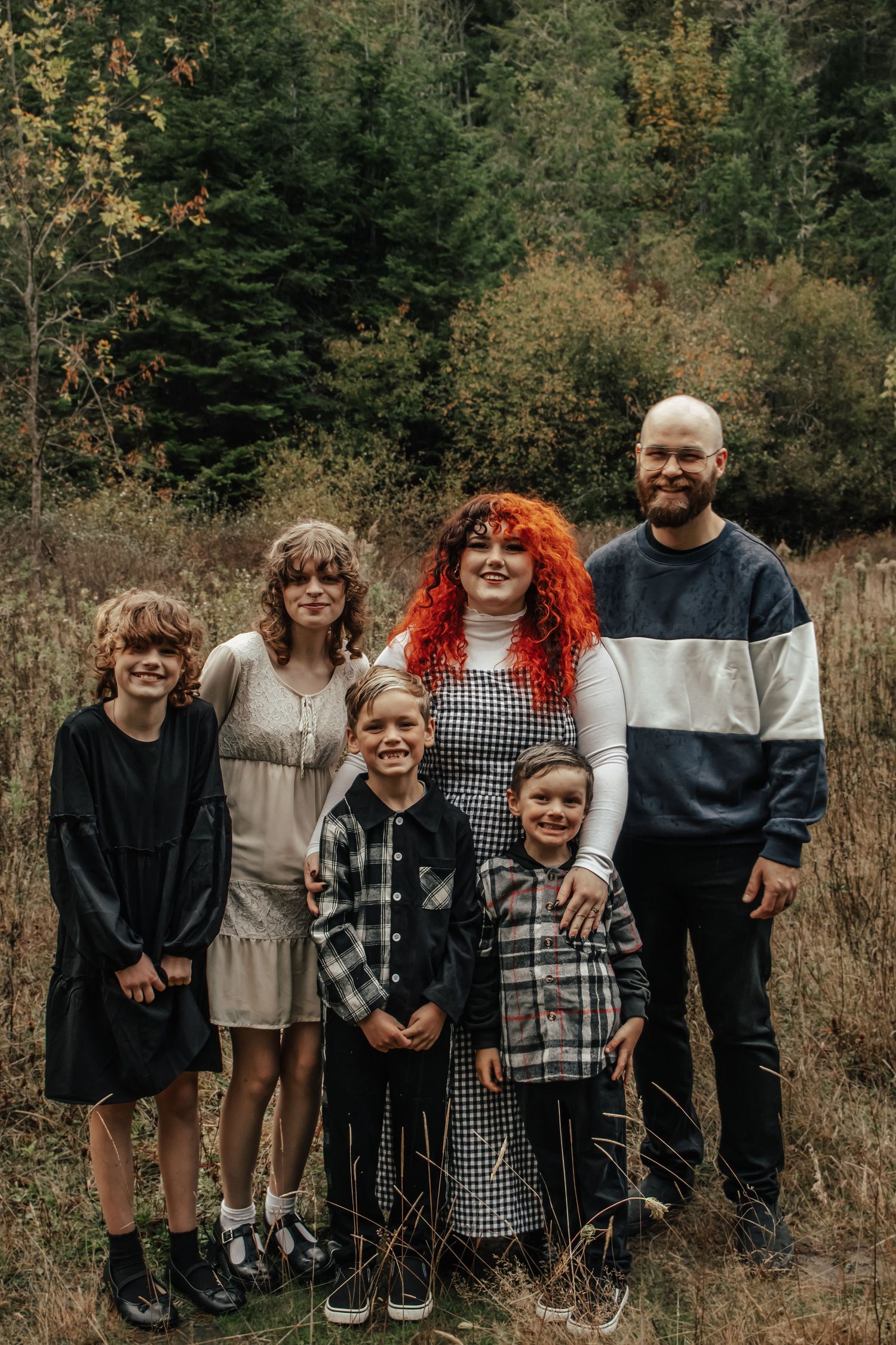 Family of six posing in an autumn meadow with evergreen trees, featuring a woman with bright red hair.