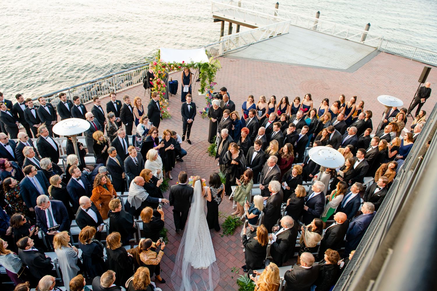 Wedding guests gather in neat rows during an outdoor ceremony with a white-dressed couple at the altar.
