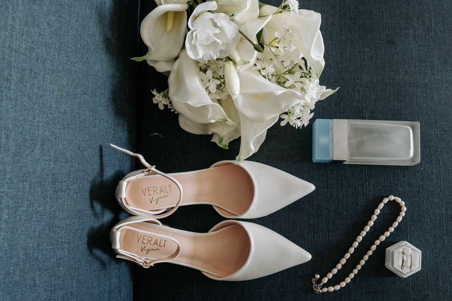 Elegant white pointed toe bridal shoes displayed with pearls and white hydrangea flowers on dark grey background.