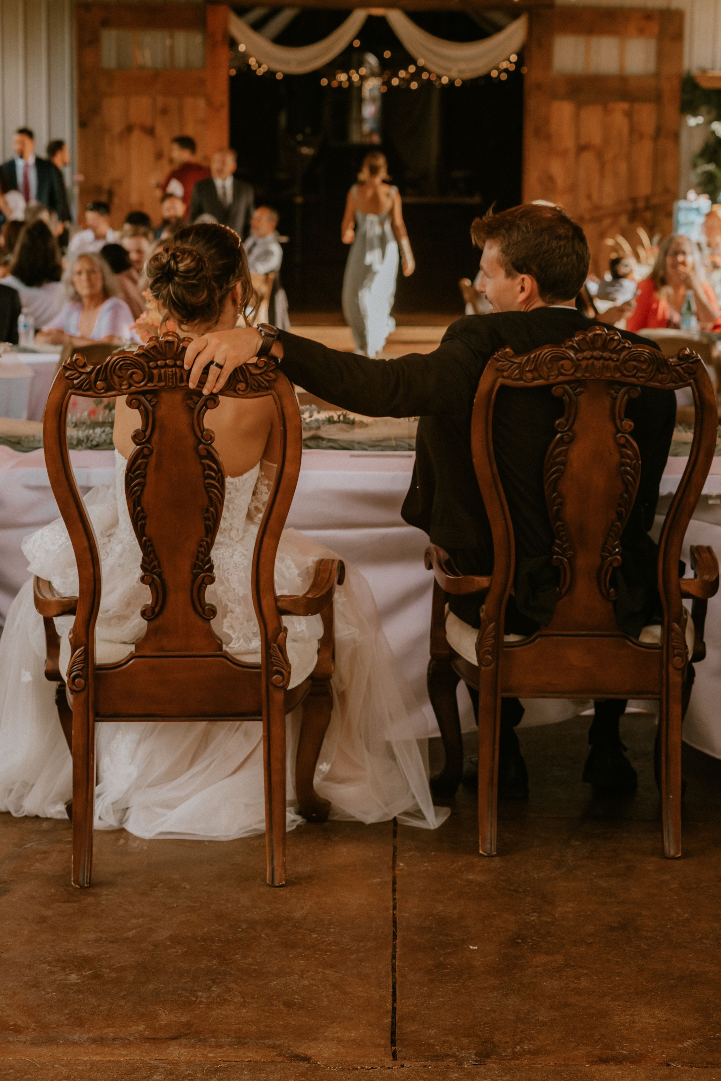 Back view of ornate vintage wooden dining chairs at a wedding reception with guests seated at tables in a rustic venue.
