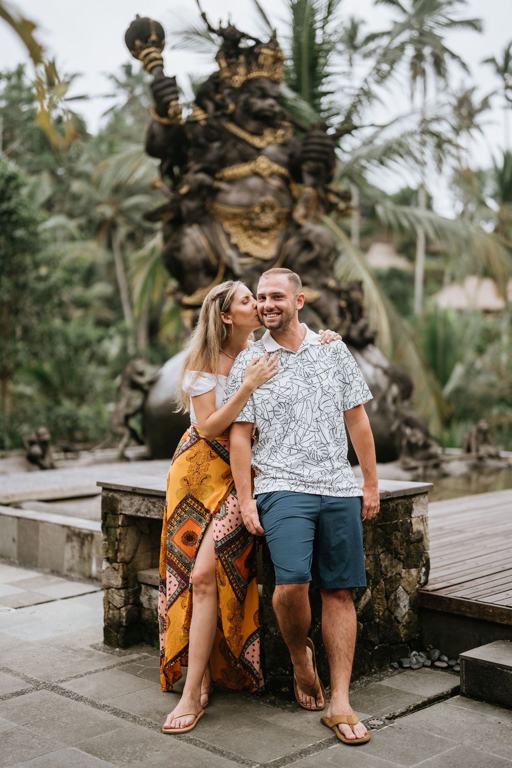 Couple in casual summer outfits sitting on wooden bench in tropical setting.